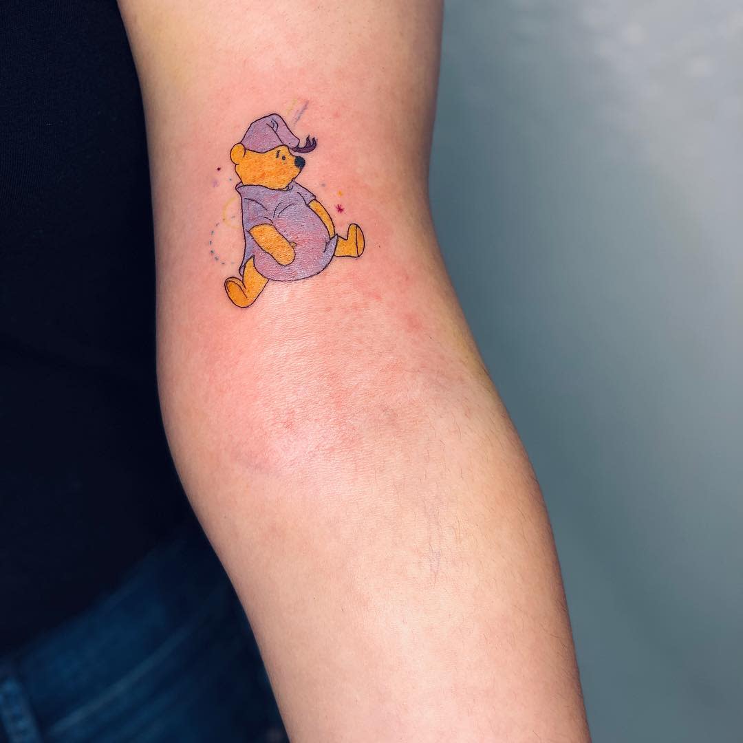 Small tattoo of Winnie the Pooh wearing a nightcap and holding a pot, with stars around him, on a person's arm